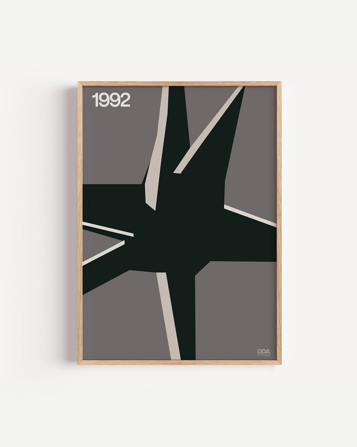 Poster 1992 di DDA in cornice rovere su parete bianca: una forma nera a raggi su fondo grigio, attraversata da lame chiare.